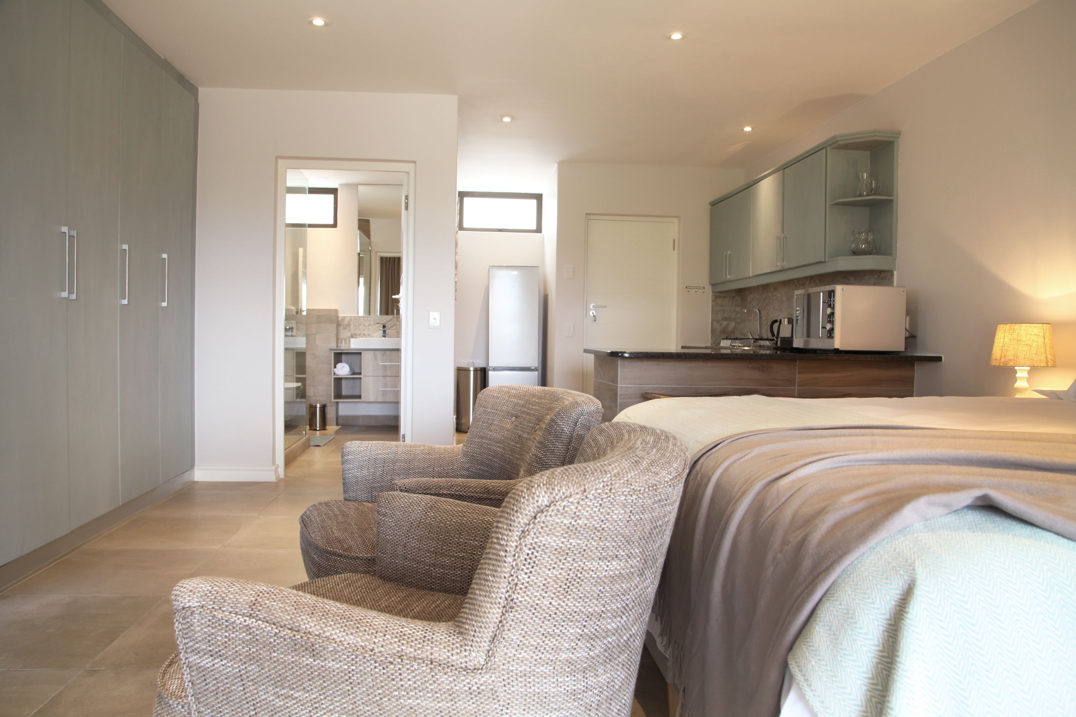 A modern bedroom with a large bed, a seating area with a chair, a kitchenette, and a bathroom visible through an open door.