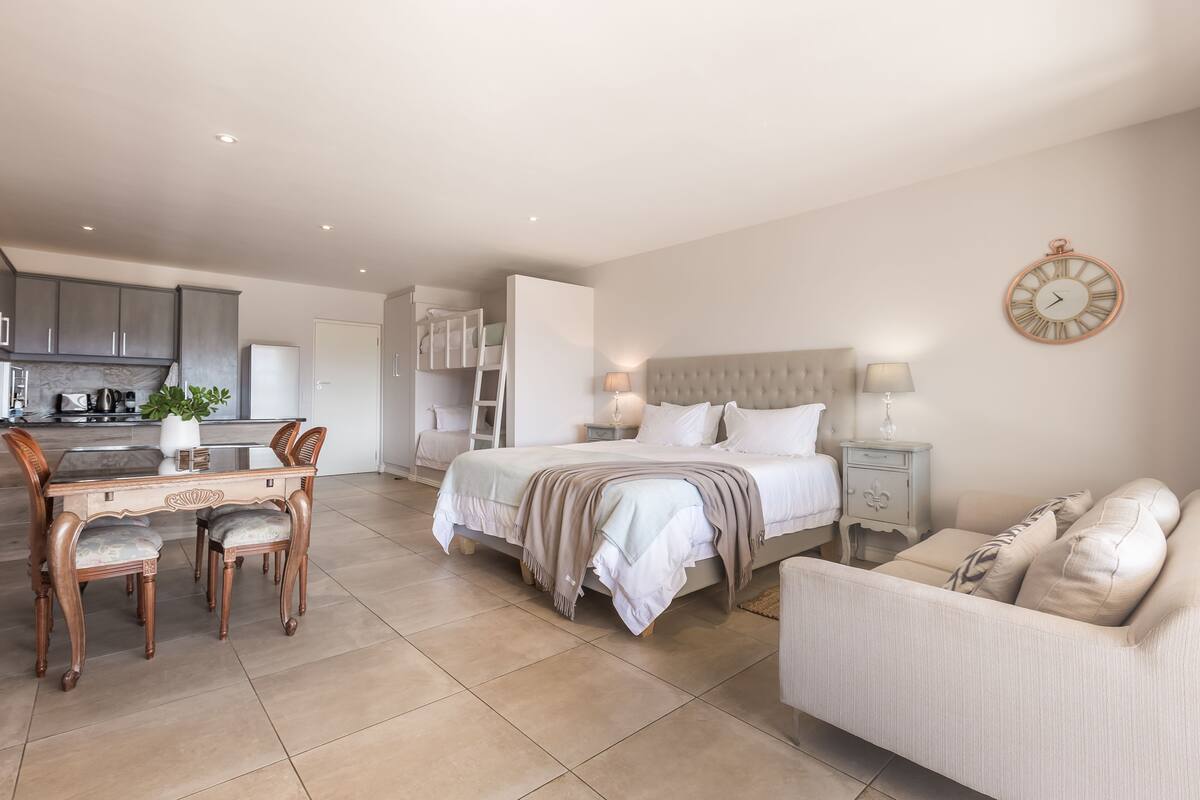 Un dormitorio moderno con cama, mesitas de noche, sofá, una mesa de comedor con sillas y una zona de cocina al fondo.