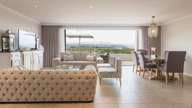 A spacious living room with a large window, a dining area with a table and chairs, and a seating area with a sofa and armchairs. - Sunrise Bay Self-Catering Accommodation (Plettenberg Bay)