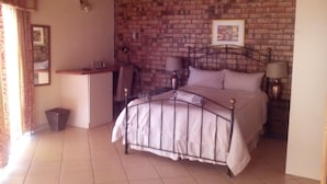 A bedroom with a brick wall, a bed with a metal frame, a bedside table with a lamp, and a small desk with a chair.