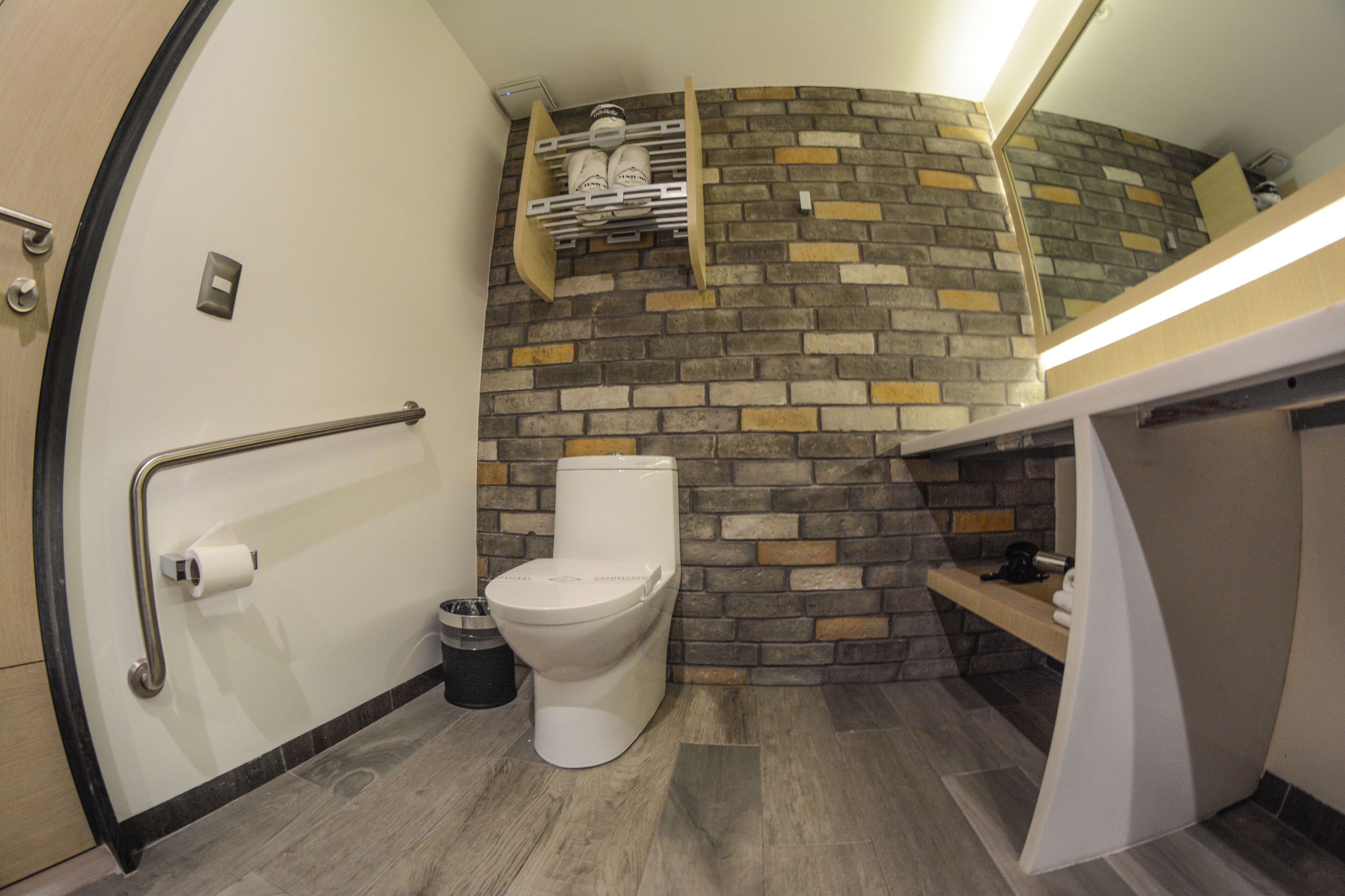 Baño compacto con inodoro, barra de apoyo, cesto de basura y repisa con artículos de aseo.