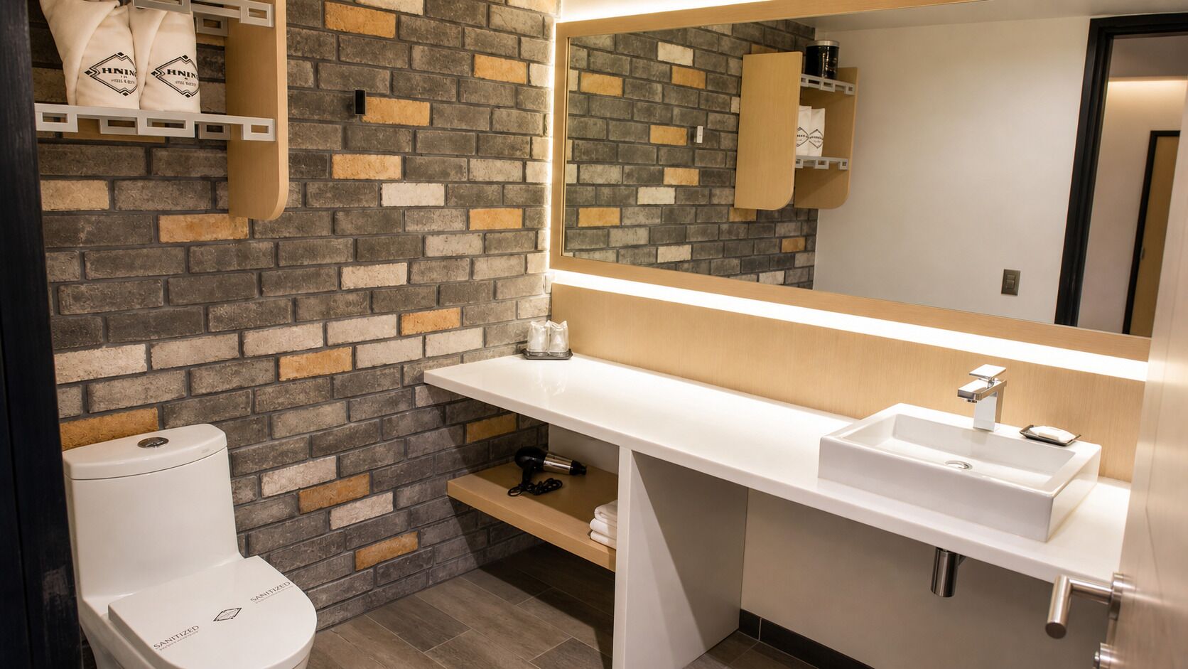 Un baño moderno con un inodoro blanco, un lavabo blanco y un espejo sobre el lavabo. La pared presenta un diseño de ladrillo.