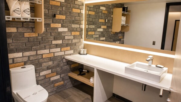 A modern bathroom with a white toilet, a white sink, and a mirror above the sink. The wall features a brick pattern.