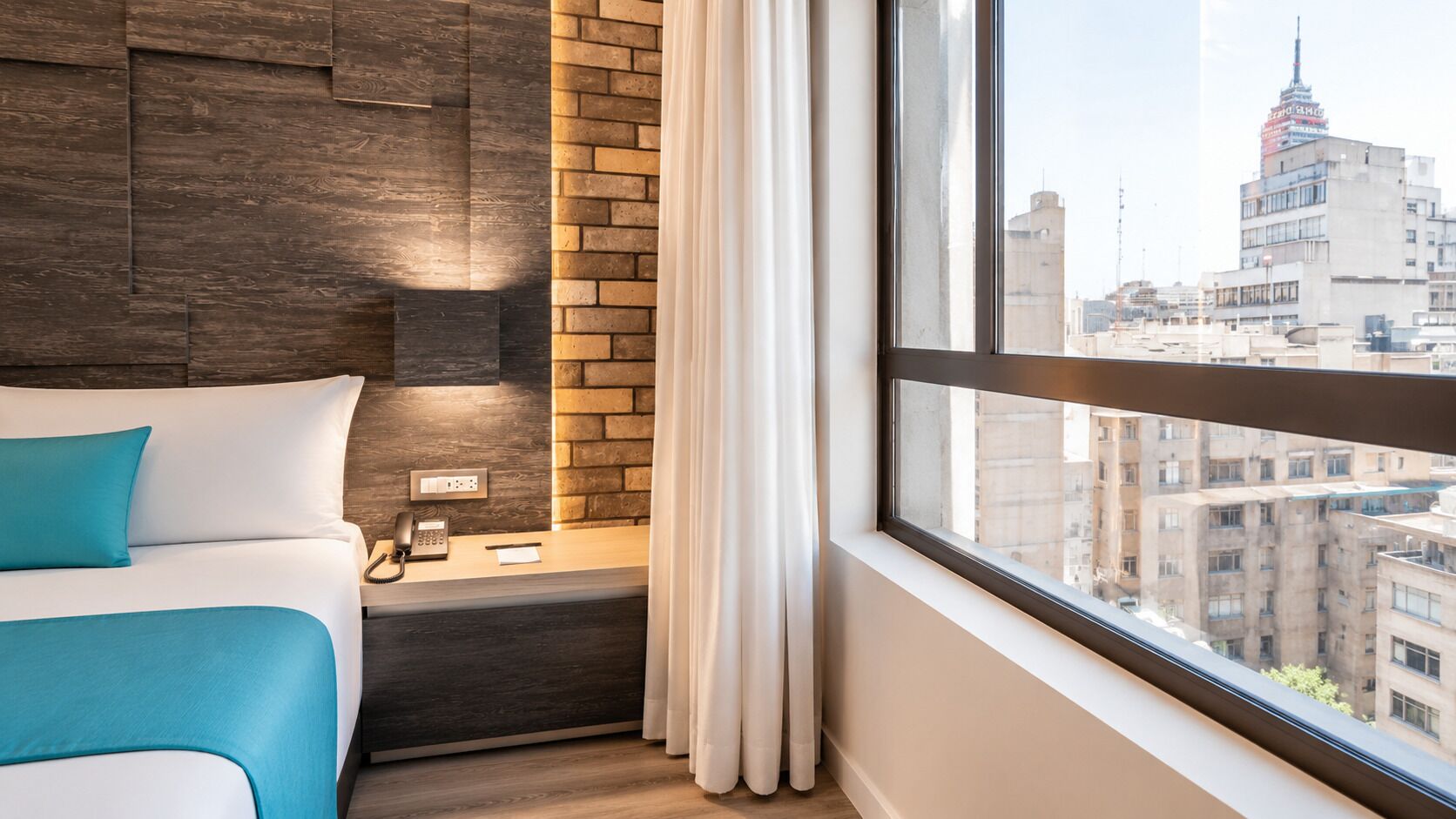 Habitación de hotel con cama, mesita de noche con teléfono, pared decorativa de ladrillo y ventana con vista a la ciudad.