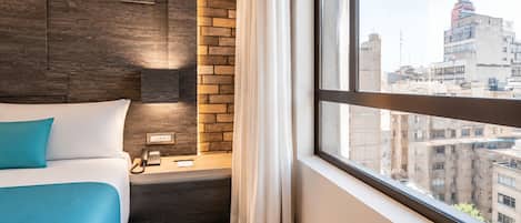 Habitación de hotel con cama, mesita de noche con teléfono, pared decorativa de ladrillo y ventana con vista a la ciudad.