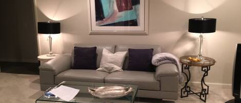 Een woonkamer met een grijze bank, twee bijzettafels met lampen, een glazen salontafel met tijdschriften, en een abstract schilderij in een lijst aan de muur.