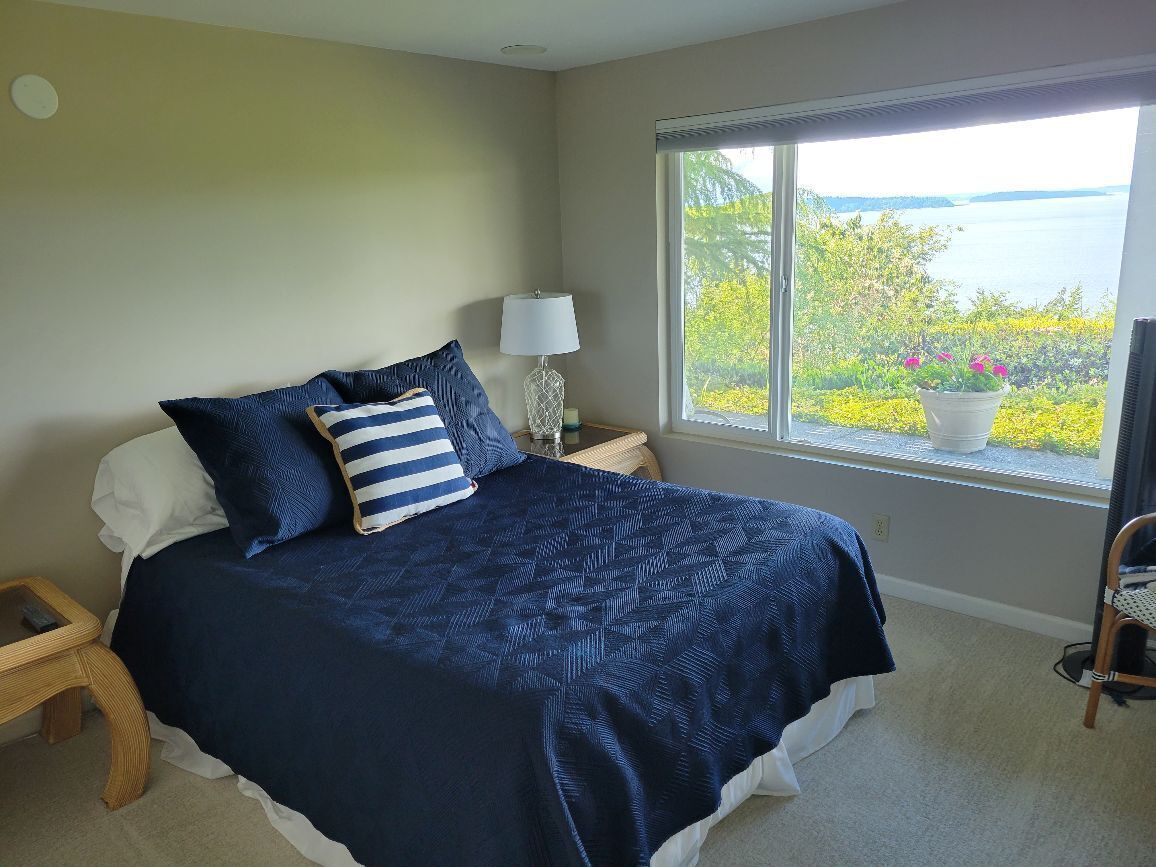 A bedroom with a large bed, a nightstand, a lamp, and a window with a view of greenery and water.