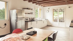 Een moderne keuken met een roestvrijstalen koelkast, een magnetron en houten kastjes. Er staat een eettafel met stoelen bij een raam dat voor natuurlijk licht zorgt.