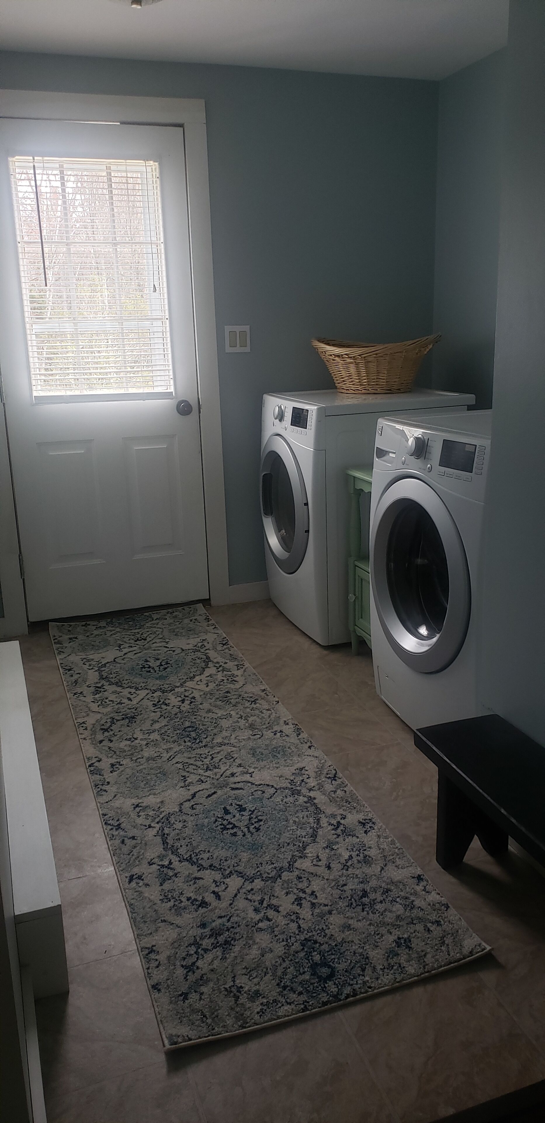 Une buanderie équipée de deux machines à laver, une porte avec une fenêtre, un tapis et un panier posé sur le dessus d’une armoire.