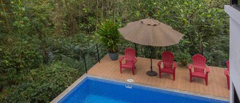 Una piscina con muelle de madera, rodeada de exuberante vegetación y sillas rojas.