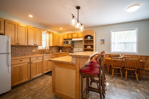 A kitchen with wooden cabinets, a central island with stools, a microwave, and a window with blinds.