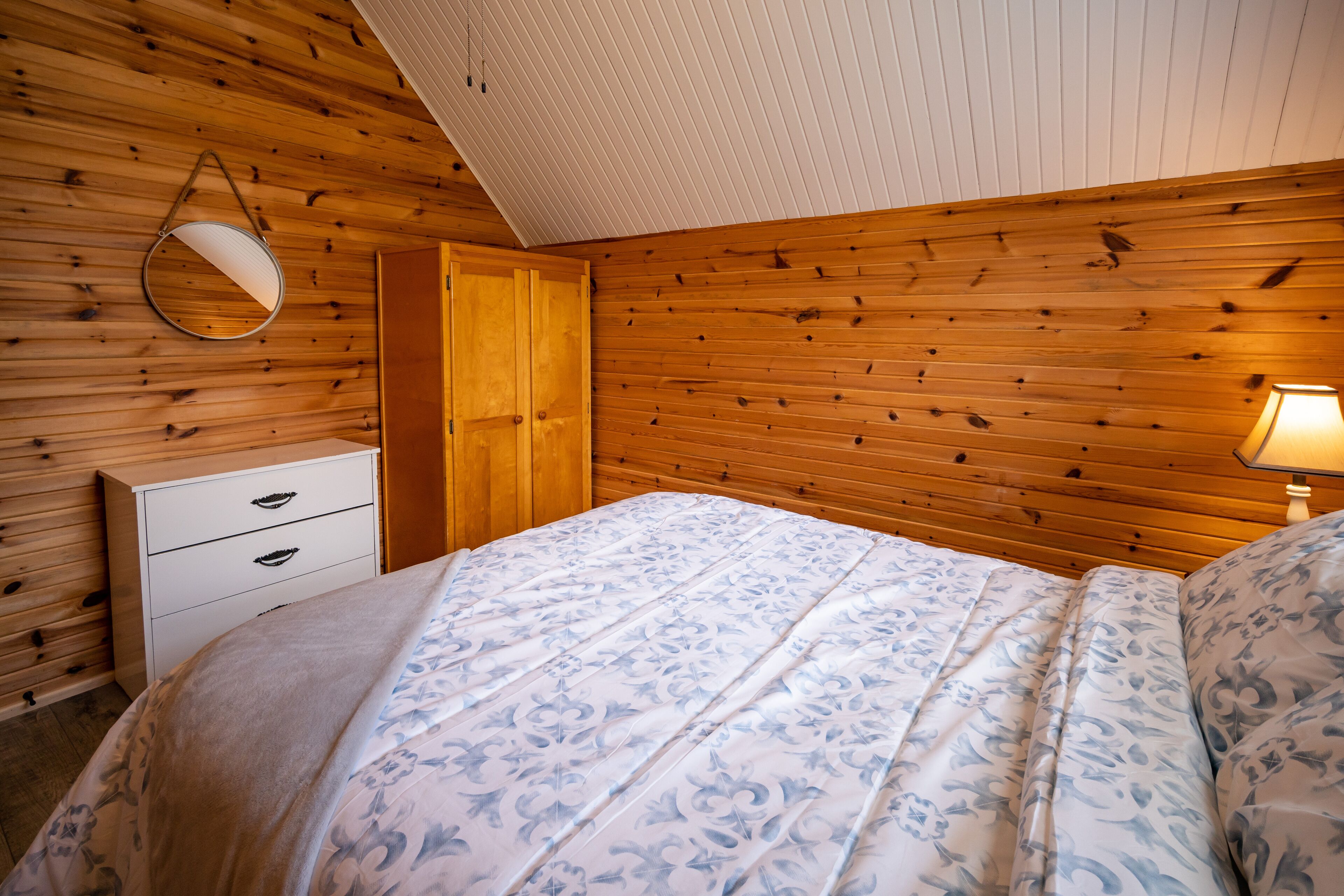 Une chambre avec un plafond en bois, un lit recouvert d’une couvre-lit à motifs floraux, une table de chevet, un miroir et une lampe.