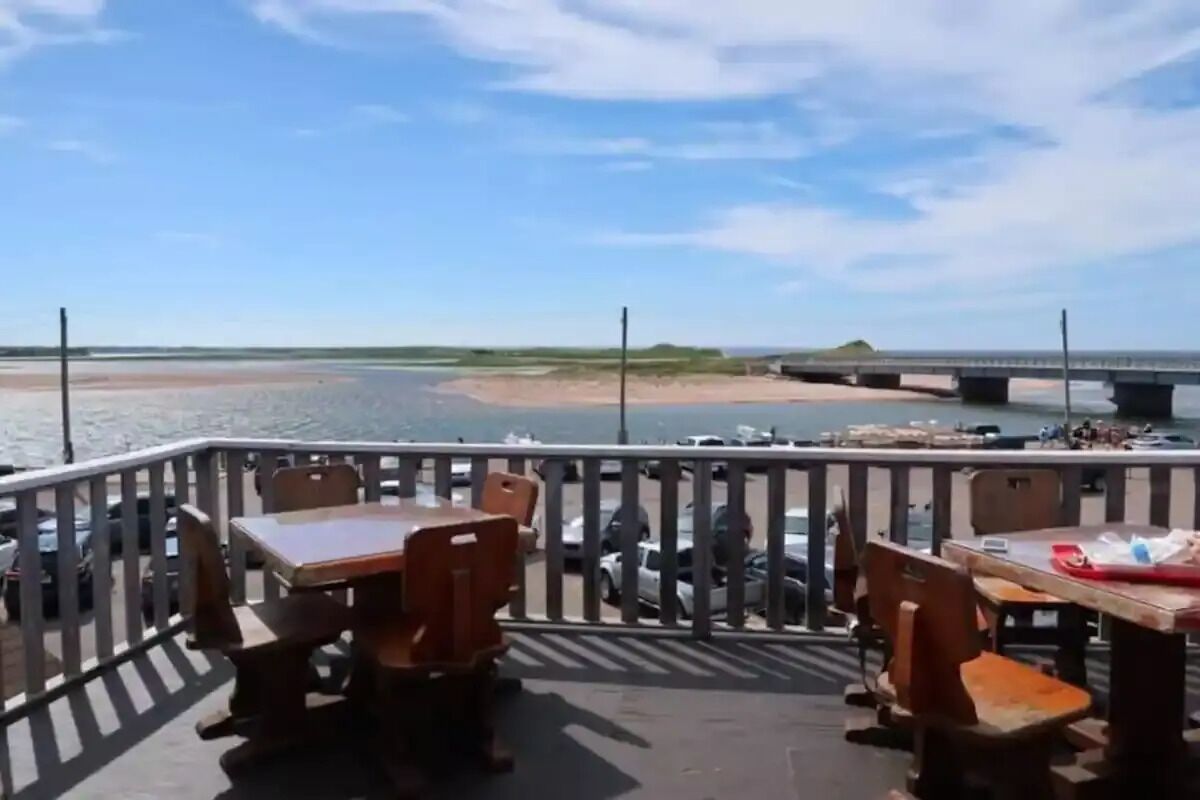 A deck with wooden tables and chairs overlooking a body of water and a bridge.