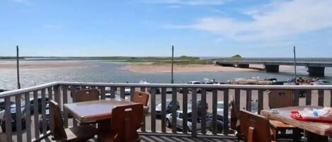 A deck with wooden tables and chairs overlooking a body of water and a bridge.