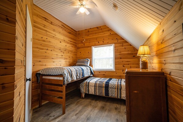 A small, cozy room with wooden walls and a sloped ceiling. It includes two beds, a small table, and a window providing natural light. - Beach, Golf, Relax & Enjoy Our Beautiful Stanhope, PEI Cottage for your vacation (Stanhope)