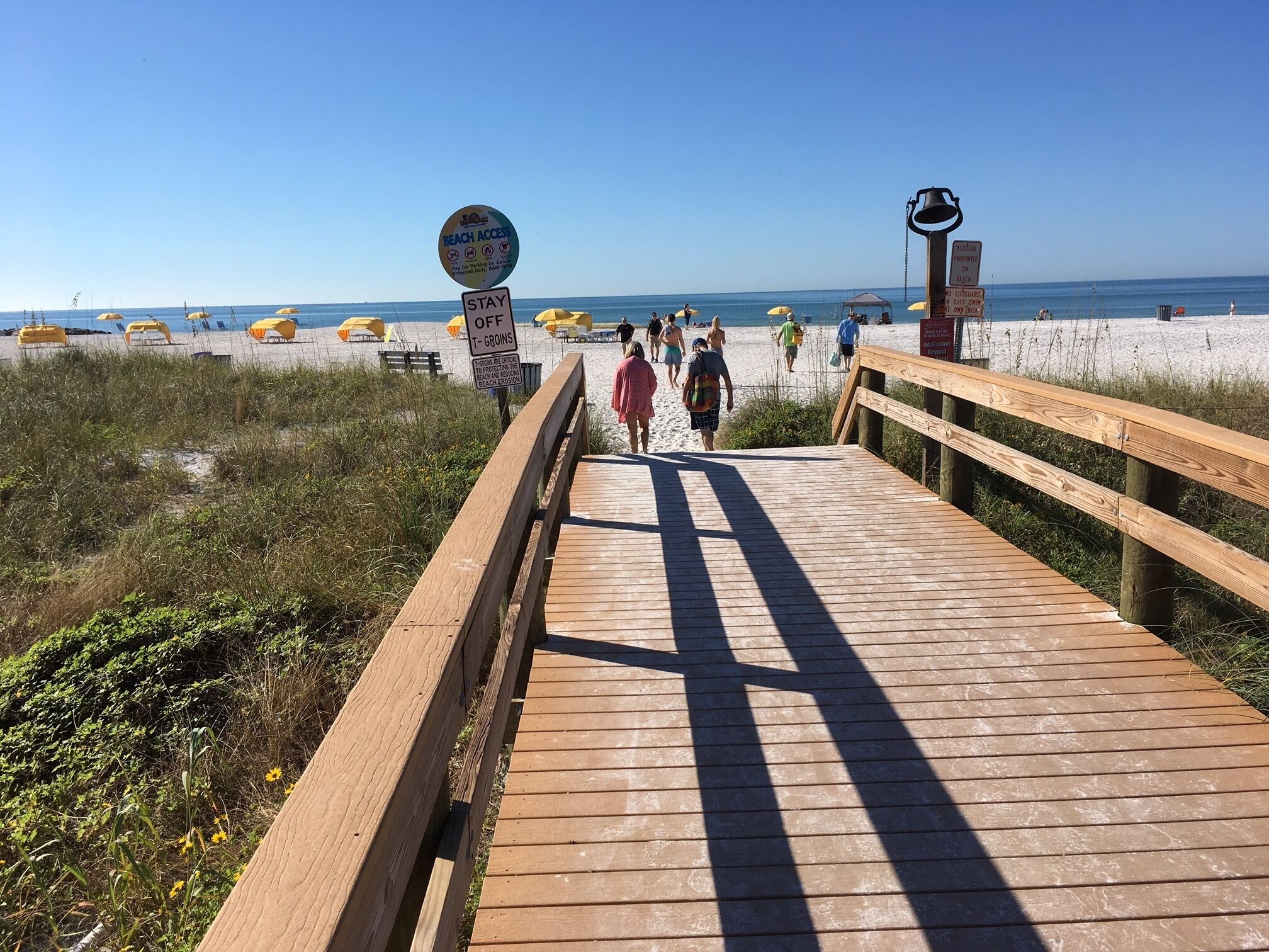Een houten looppad dat naar een strand leidt, met parasols en mensen die erover lopen.