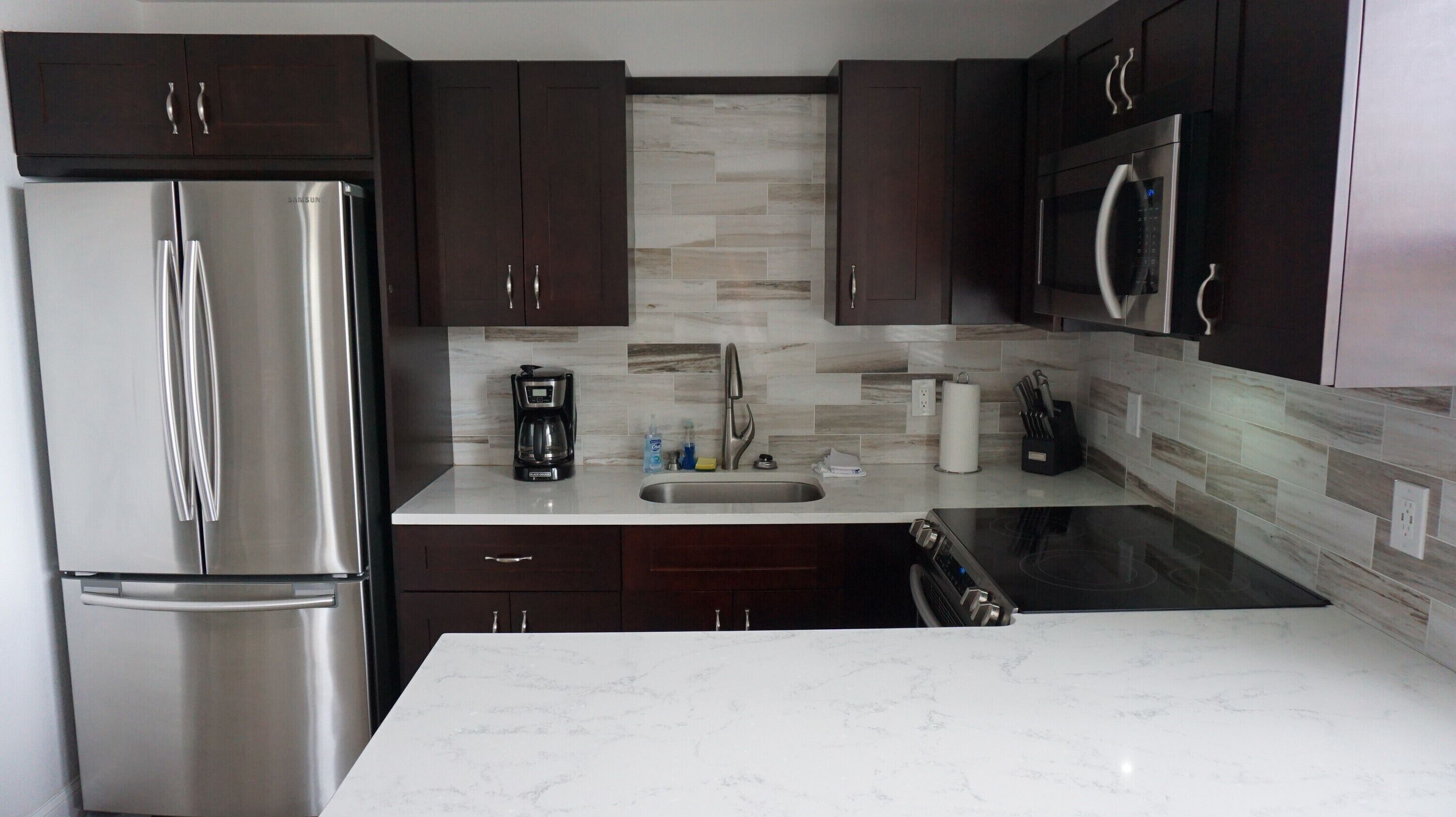 A modern kitchen with dark wooden cabinets, a stainless steel refrigerator, a built-in microwave, and a marble countertop.