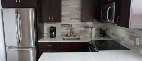 A modern kitchen with dark wooden cabinets, a stainless steel refrigerator, a built-in microwave, and a marble countertop.