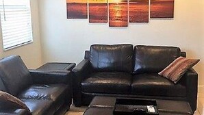 A living room with a black leather sofa, a matching ottoman, and a coffee table. A large wall-mounted painting of a sunset is visible. - Beach, Pool, Restaurants, Paradise ~ FN 803 (St Pete Beach)