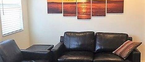 Een woonkamer met een zwarte leren bank, een bijpassend voetenbankje en een salontafel. Een groot schilderij van een zonsondergang hangt aan de muur.