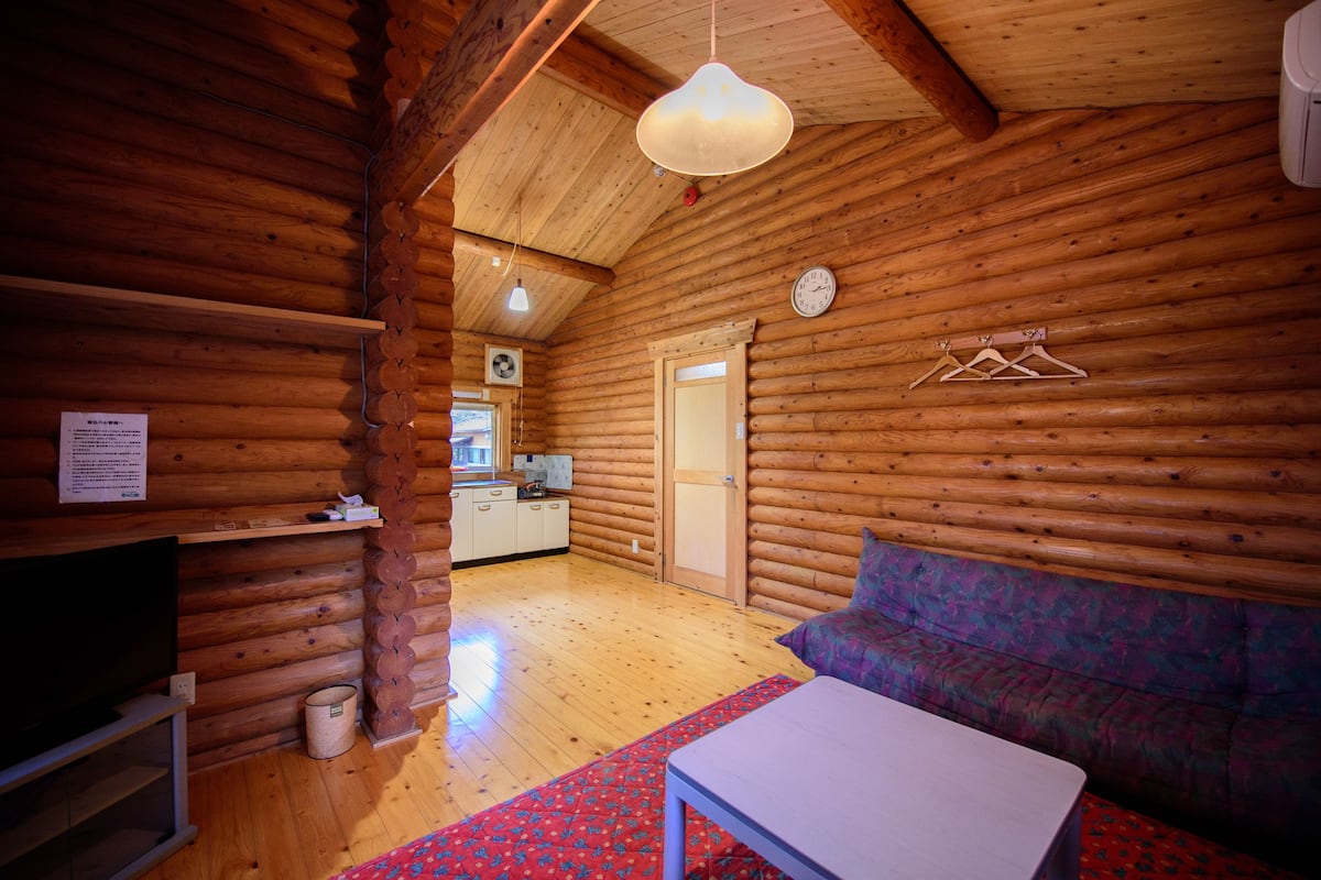 Ein Holzhaus-Interieur mit Holzboden, einer Couch, einem Tisch, einer hängenden Leuchte, einer Uhr und einem durch eine offene Tür einsehbarer Küchenbereich.