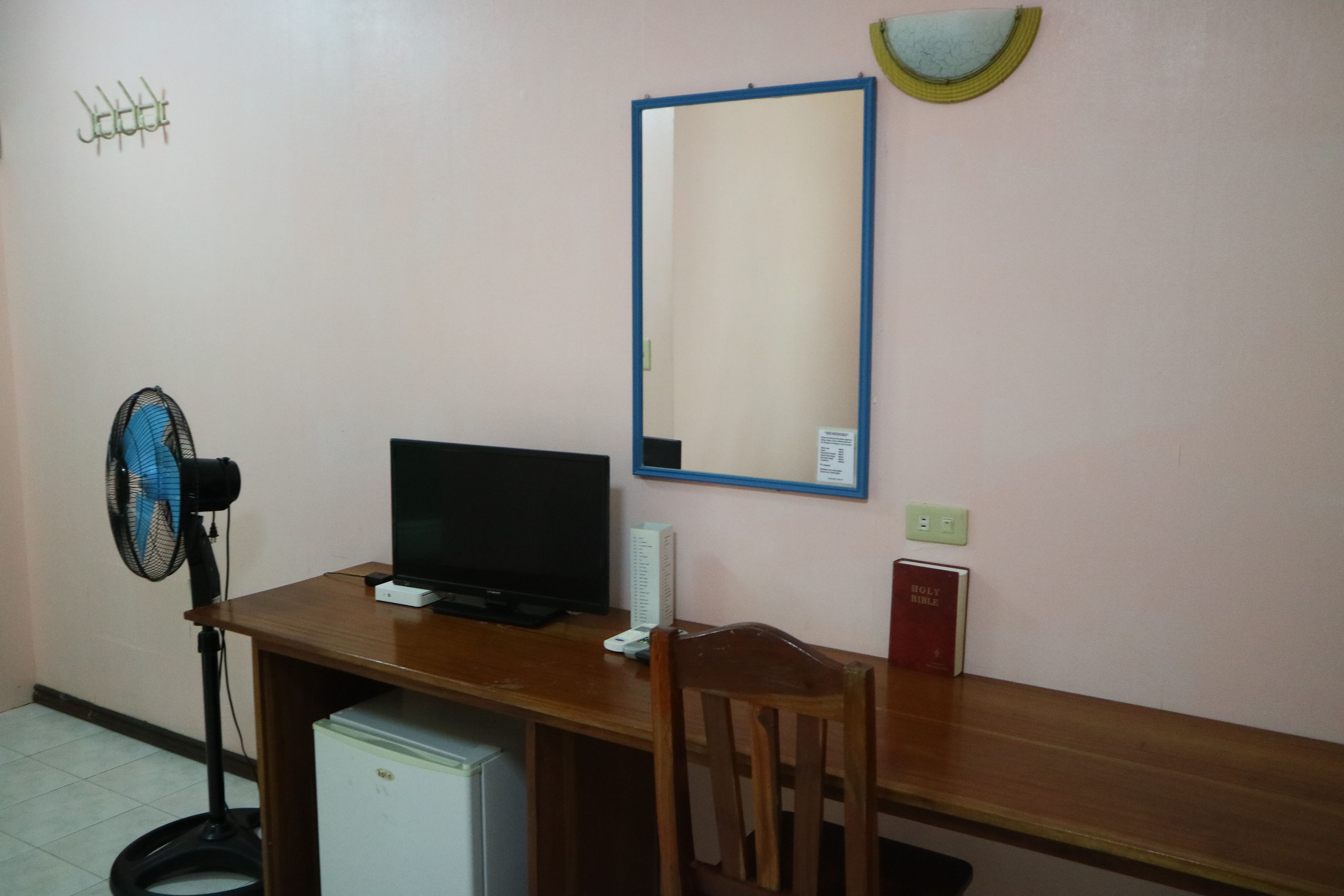 A small room with a wooden desk, a black computer monitor, a fan, a small refrigerator, and a chair.