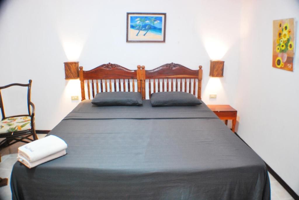 A single bed with a wooden headboard, two pillows, a black bedspread, a chair with a floral cushion, a small table with a lamp, and a painting on the wall.