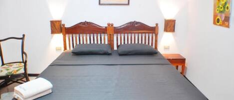 A single bed with a wooden headboard, two pillows, a black bedspread, a chair with a floral cushion, a small table with a lamp, and a painting on the wall.