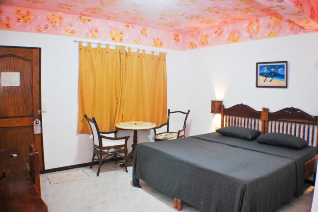 Una habitación con una cama, una mesa, dos sillas, una puerta y una pared decorada con un cuadro.