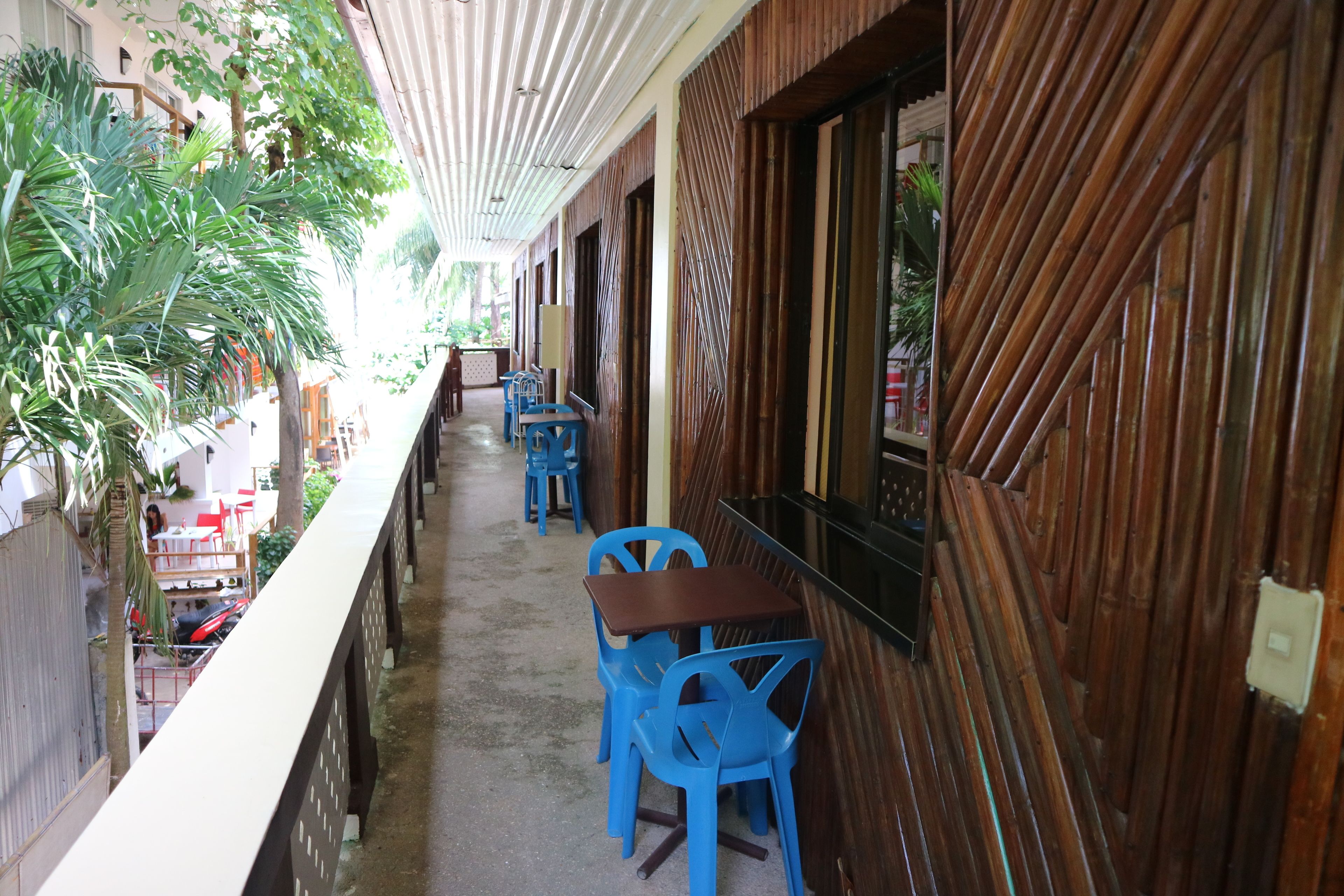 Un espace extérieur abrité, avec des chaises et des tables en plastique bleu, des murs en bois et une vue sur la rue.
