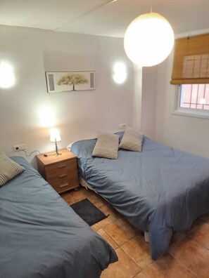 A small, well-lit bedroom with two single beds, a bedside table with a lamp, a small dresser, and a window with a view.