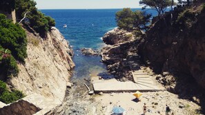 A rocky coastal area with a clear view of the sea, a small boat, and a paved path leading to a beach.