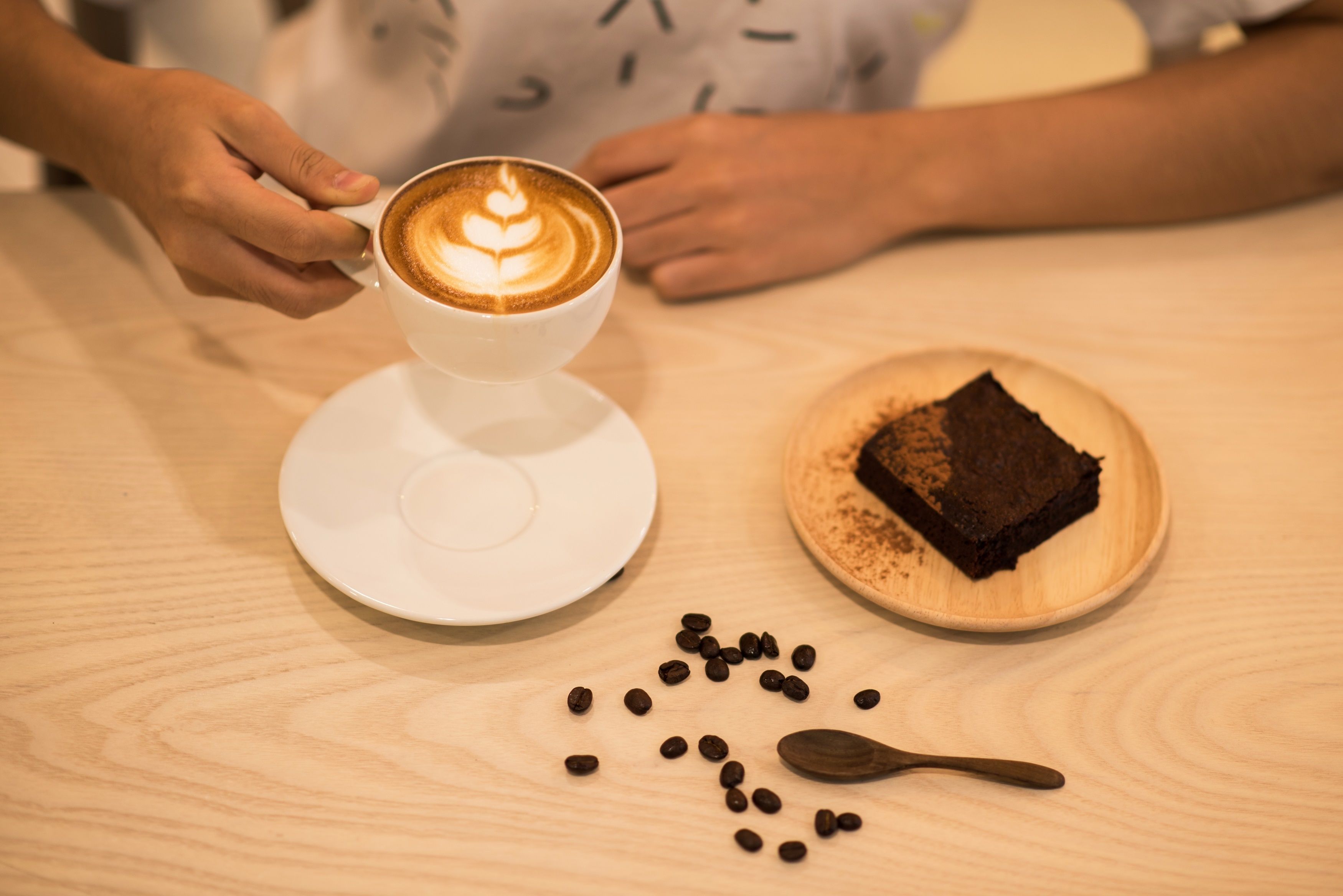 A person holding a cup of coffee with latte art, a plate of brownies, and scattered coffee beans on a wooden table.