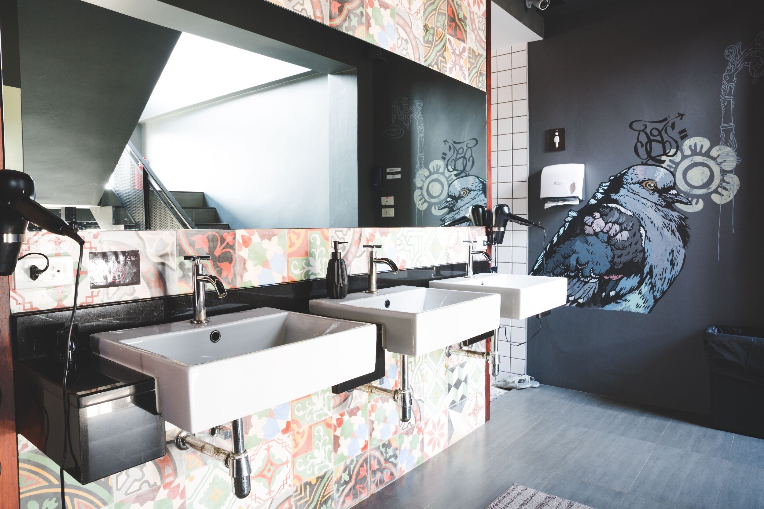 Un baño moderno con tres lavamanos, un espejo grande y un mural de un pájaro en la pared.