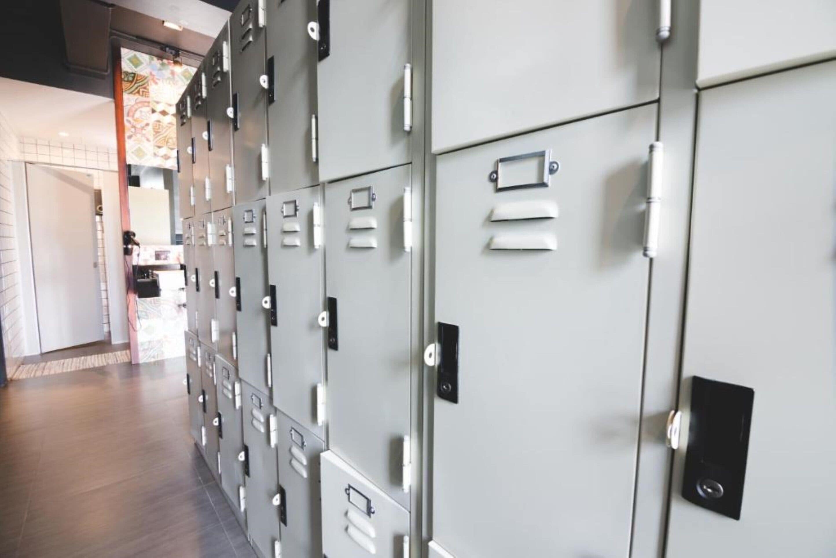 A row of grey lockers with white handles and a black lock plate, in a hallway with a tiled floor.