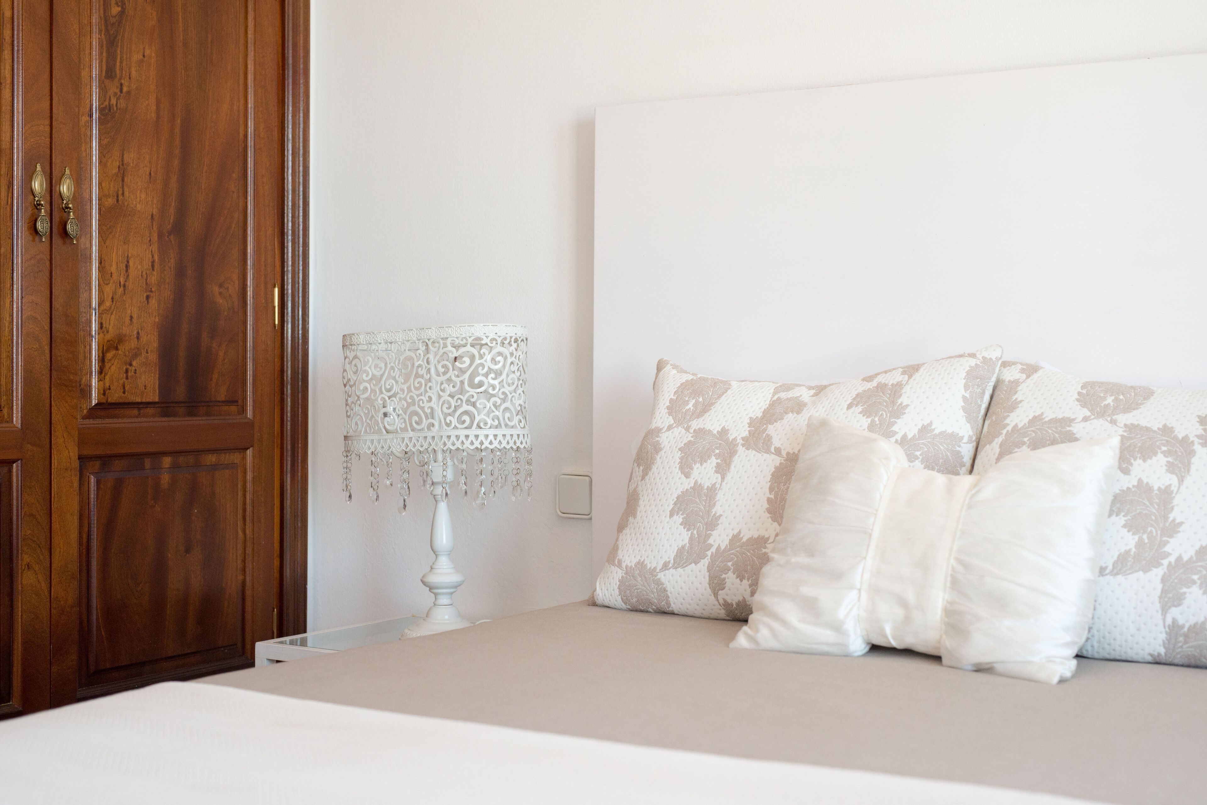 A bedroom with a wooden wardrobe, a white bedside table with a patterned lamp, and a bed with patterned pillows.