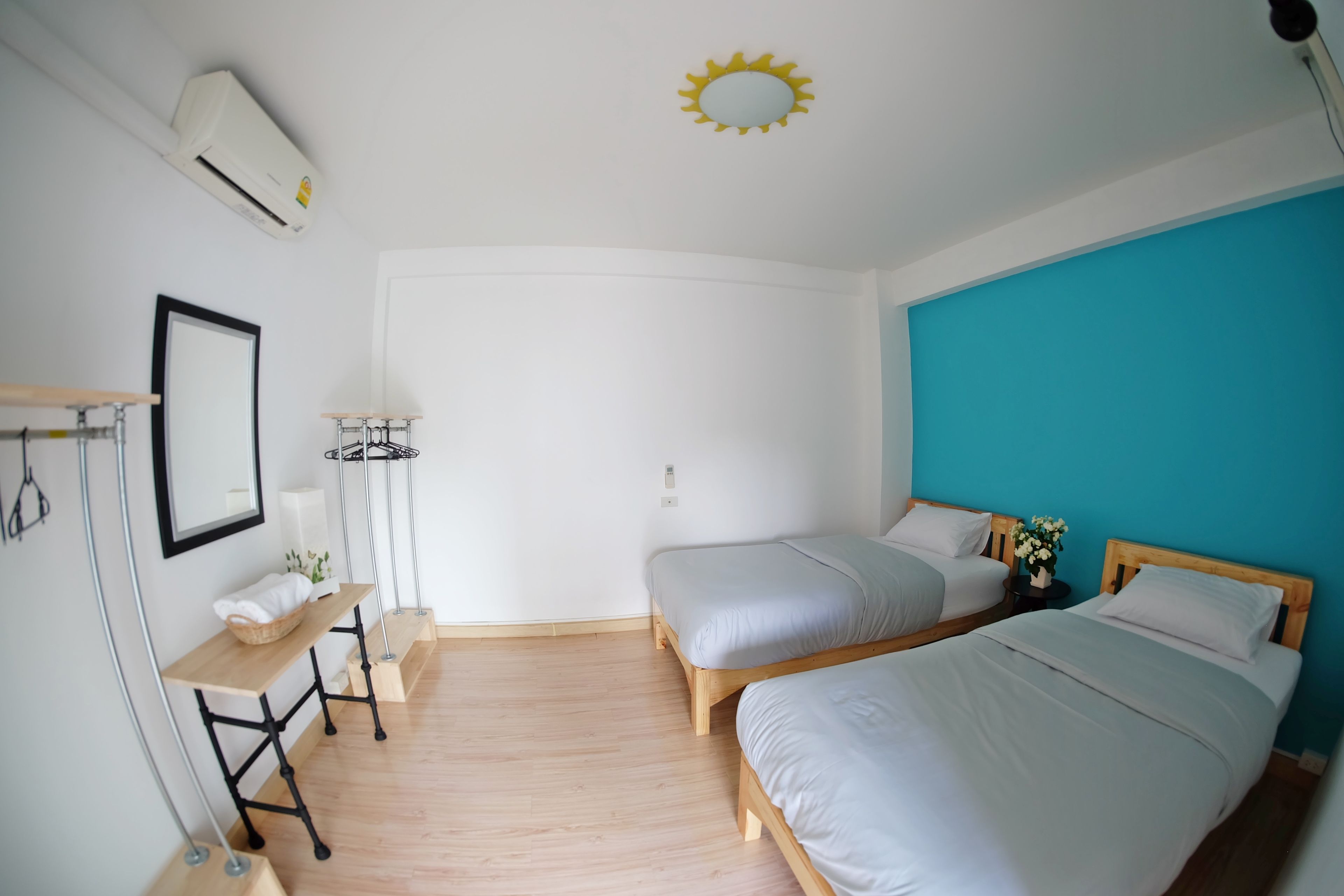 A room with two beds, a desk, a chair, a small table, a wall-mounted air conditioner, a ceiling light, and a wall-mounted mirror.