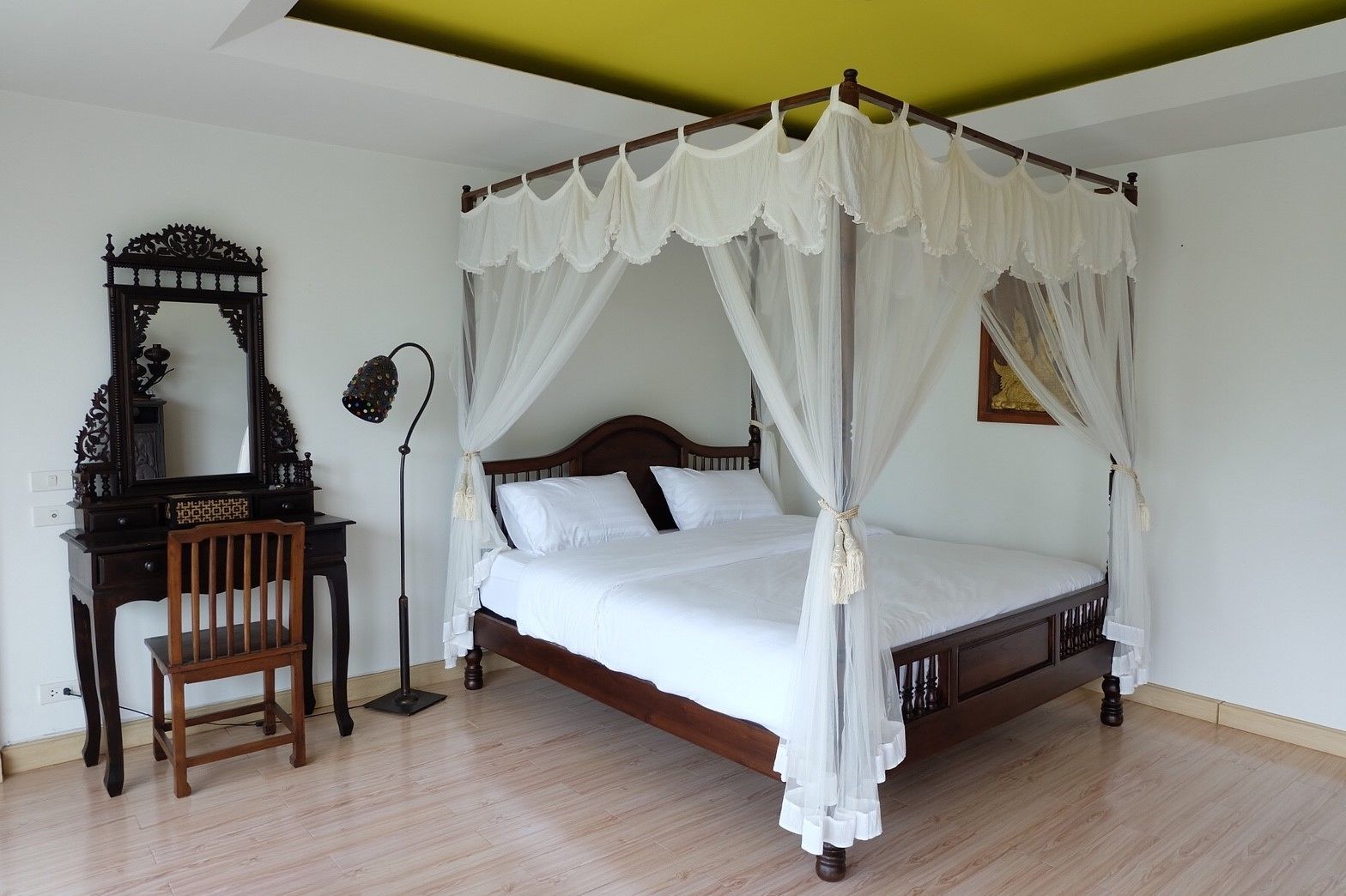 A four-poster canopy bed with white bedding, a wooden nightstand with a mirror, and a desk with a chair.