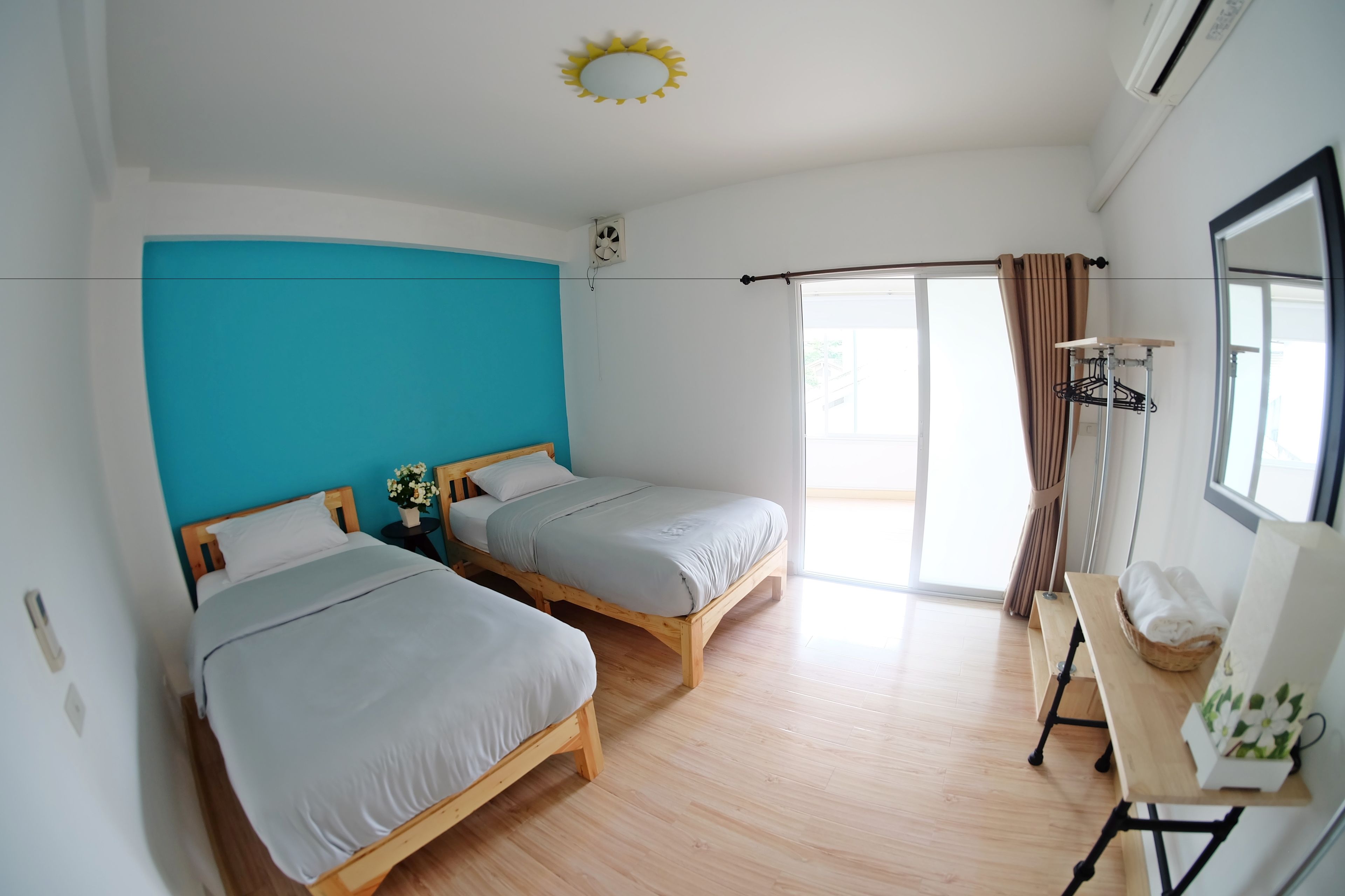 Une chambre avec deux lits en bois, une petite table, un ventilateur et une fenêtre avec des rideaux.