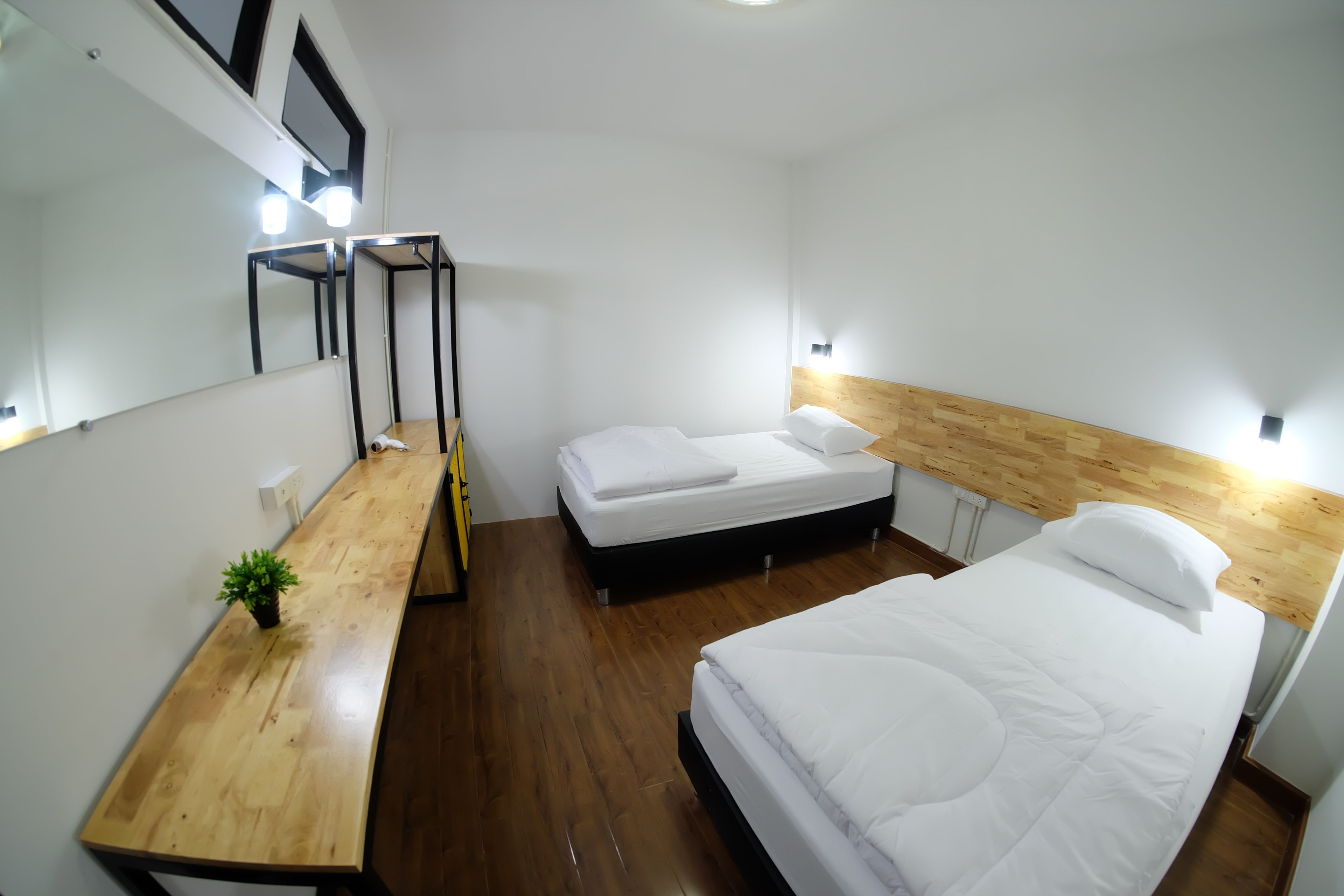 A hotel room with a wooden floor, a bed with white bedding, a wooden headboard, a bedside table with a plant, and a mirror with lights.
