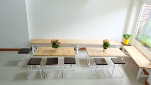 Habitación limpia y moderna con mesas y bancos de madera, plantas en macetas y un ventanal que proporciona luz natural.