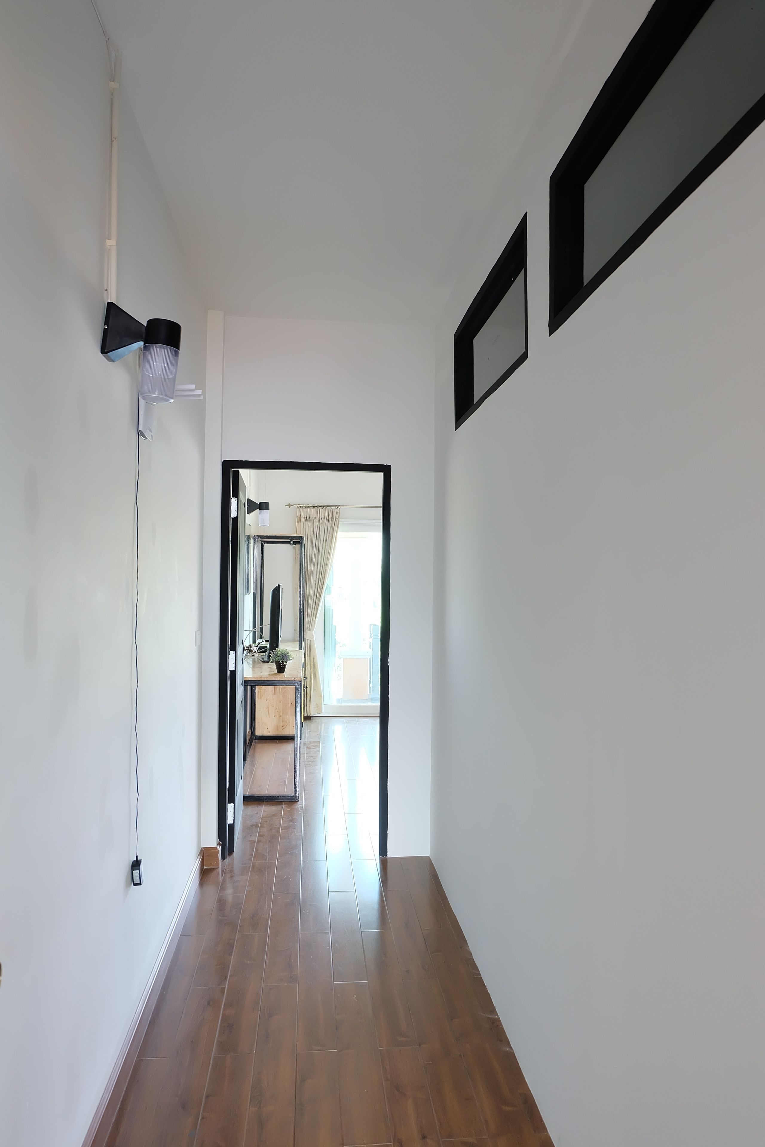 A hallway with a wooden floor, a black-framed mirror, and a light fixture on the left.