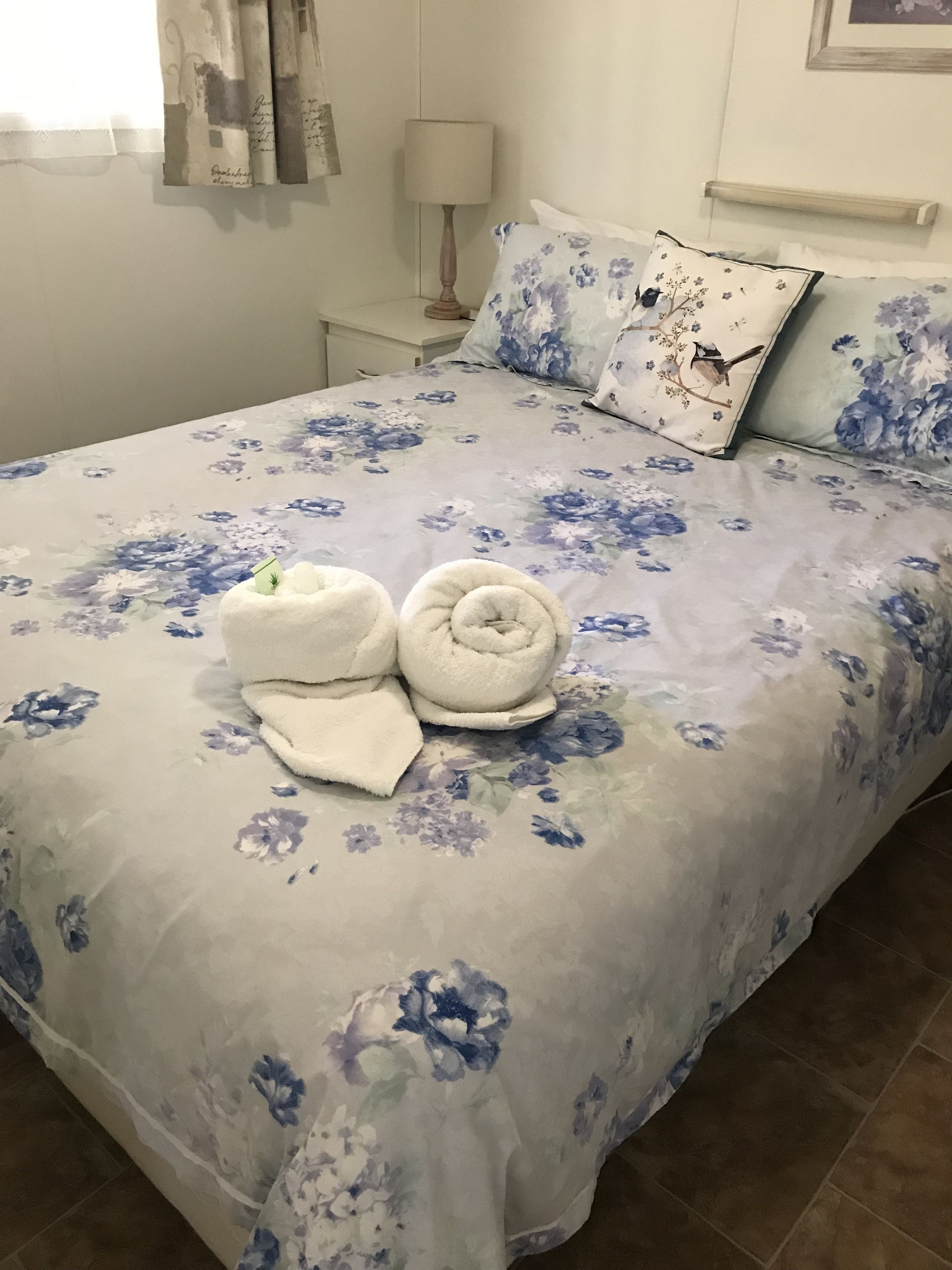 A bed with a floral bedspread, two rolled towels, and a nightstand with a lamp.