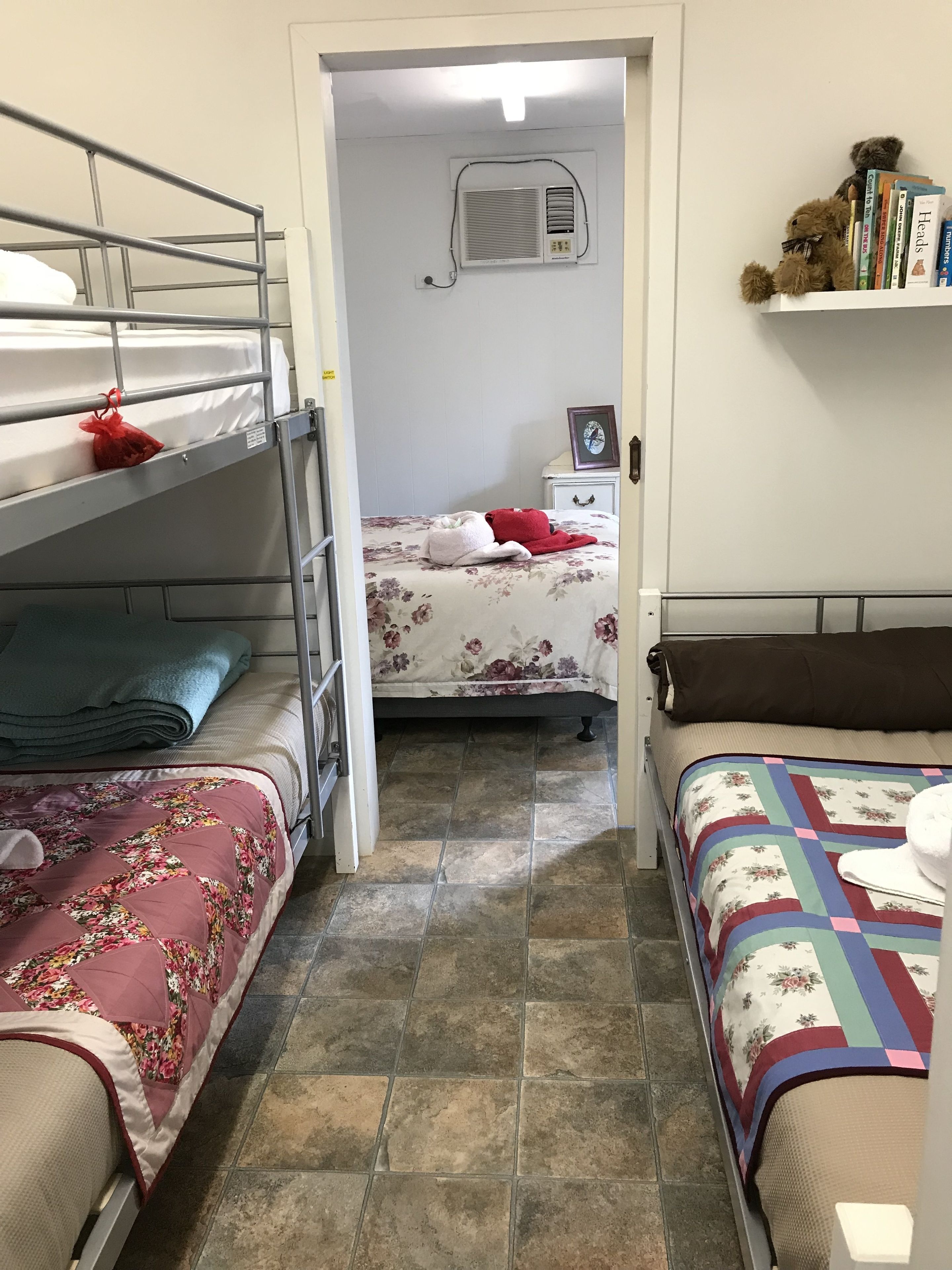 Une chambre de dortoir avec des lits superposés, un lit simple, une table de chevet avec une lampe, un climatiseur fixé au mur et une étagère avec des livres et un ours en peluche.