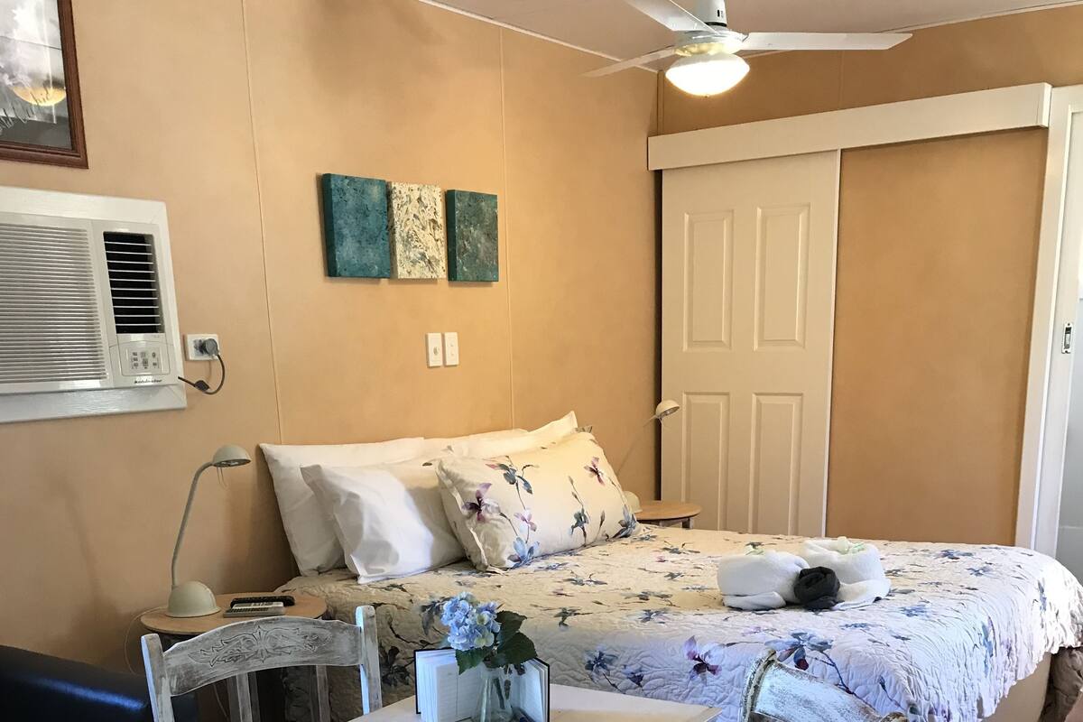 A room with a bed, a table, and a chair. There is a ceiling fan, a lamp, and a wall-mounted air conditioner.