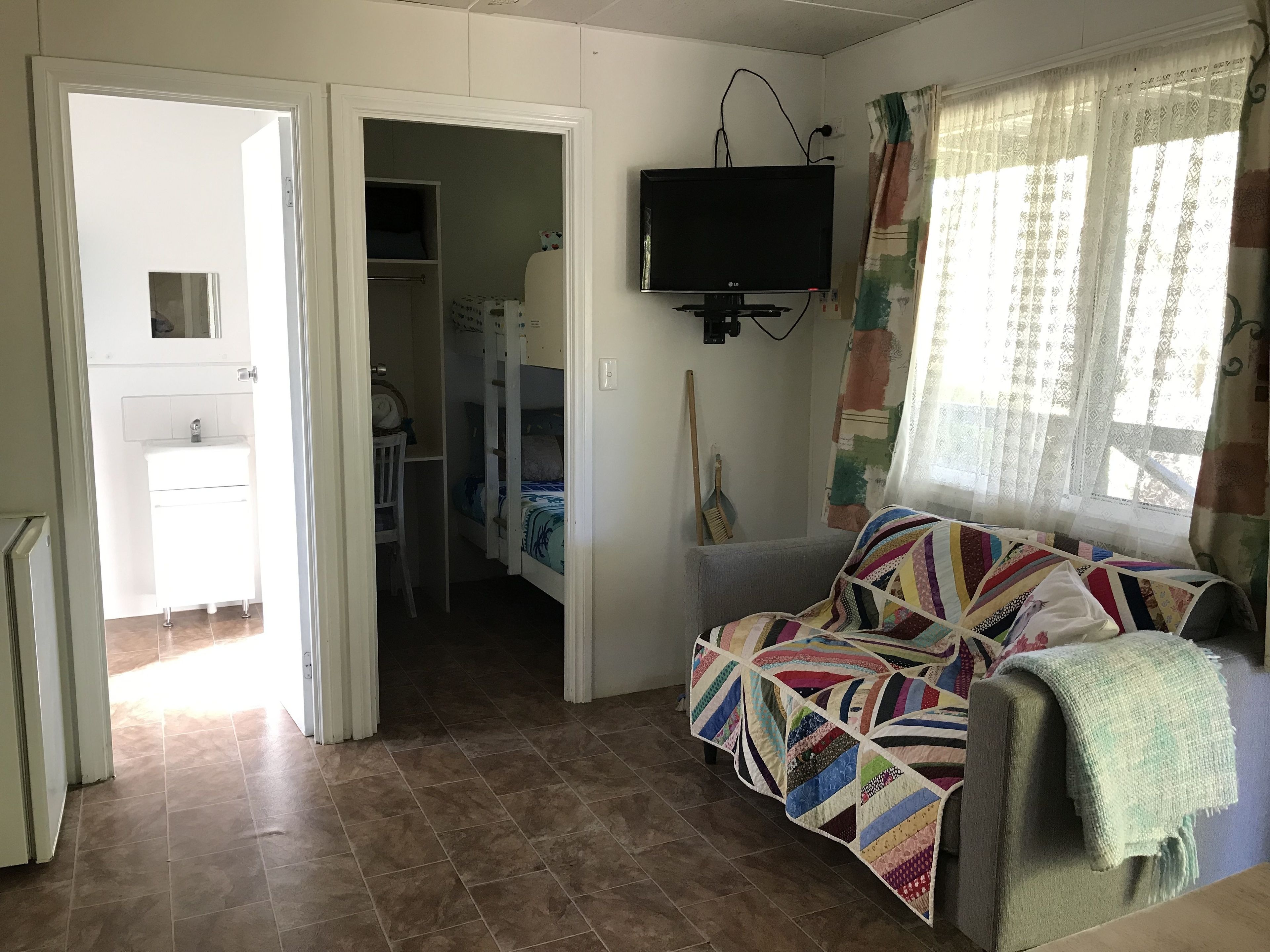 A room with a sofa, a television, a window with curtains, a door leading to a bathroom, and a tiled floor.