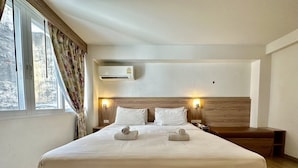 A hotel room with a large bed, two bedside lamps, an air conditioning unit, and a window with curtains. - Dinsomon (Bangkok)
