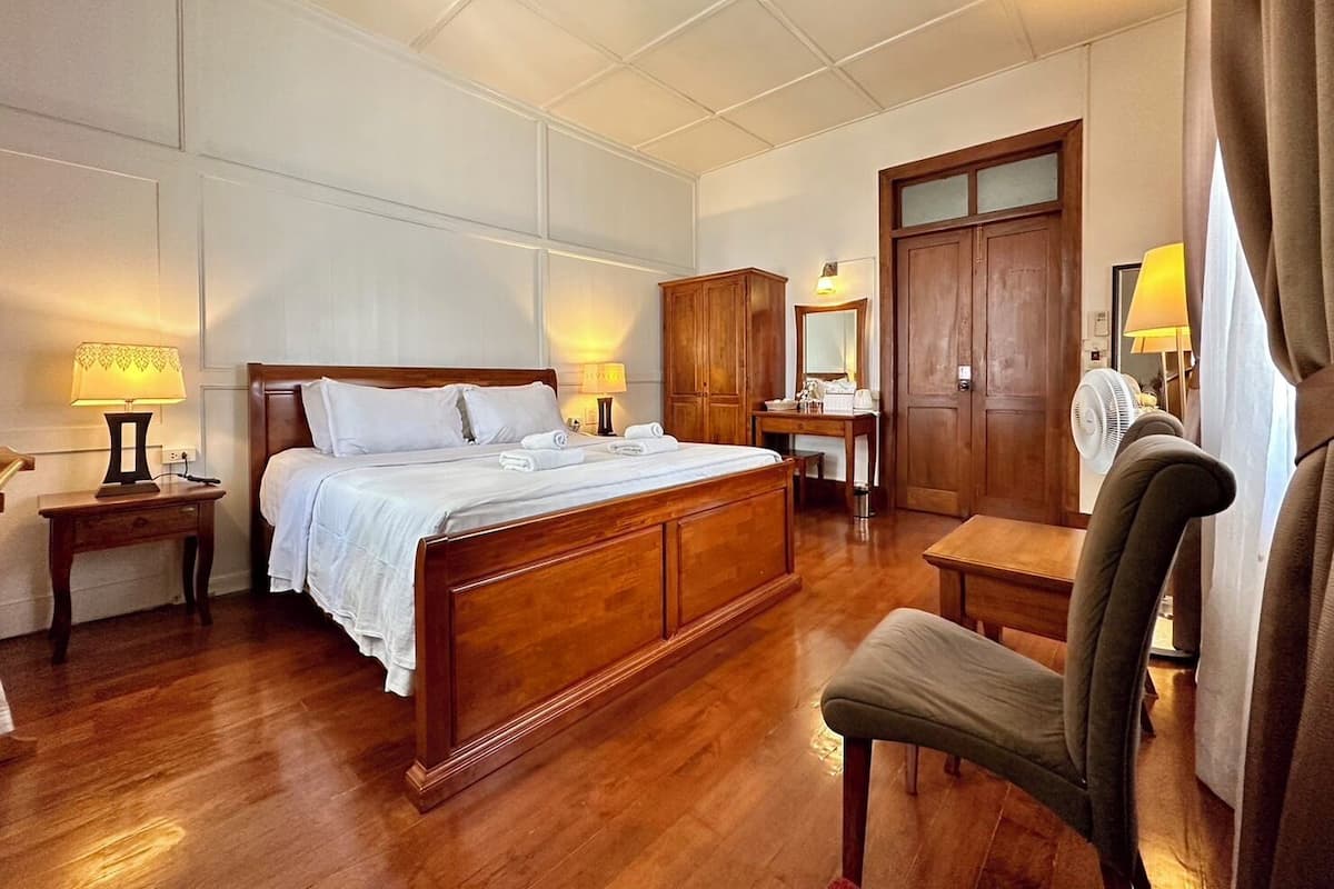 A hotel room with a large bed, wooden headboard, bedside tables with lamps, a desk with a mirror, a chair, and a wardrobe.