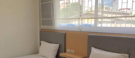 A hotel room with two single beds, a small bedside table, a window with blinds, and a view of buildings outside.