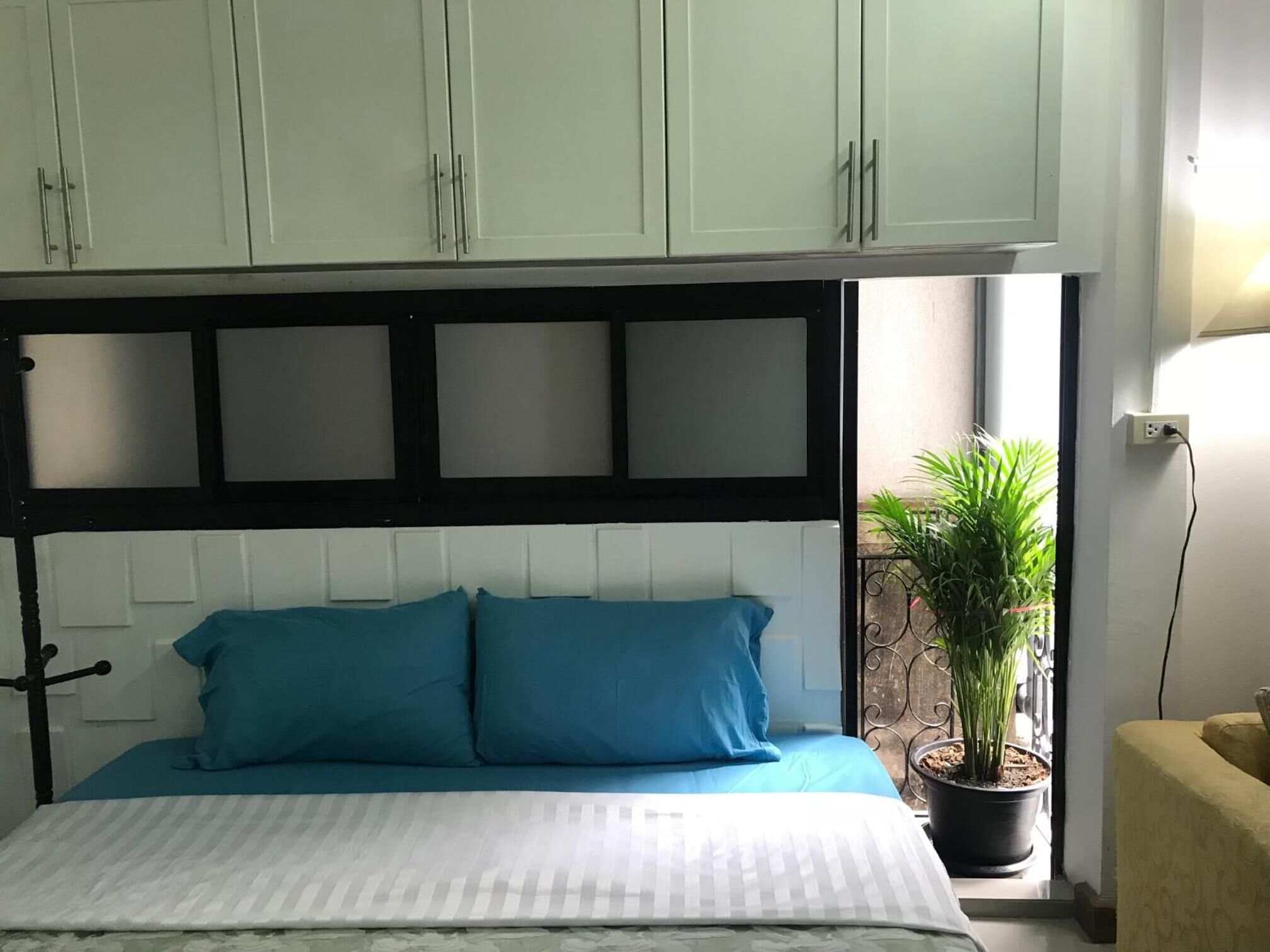 Uma cama com colcha listrada em branco e azul, cabeceira preta e duas almofadas azuis. Há uma planta em vaso num parapeito de janela à direita.